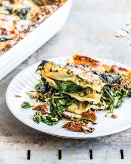 Ein weißer Teller mit Spinat Lasagne daneben Besteck und eine Auflaufform
