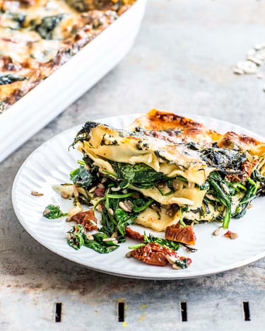 Ein weißer Teller mit Spinat Lasagne daneben Besteck und eine Auflaufform