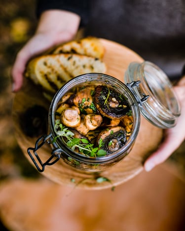 Glas mit marinierten Pilzen auf Holzbrett mit Ciabatta daneben.