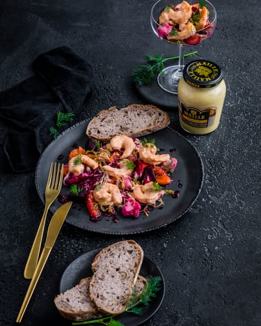 Auf dunklen grauen Tellern ist ein Salat mit Garnelen  und Brot angerichtet. EinTeil des Salates ist in einem Cocktailglas, zwei Scheiben Brot und ein Glas Mayonnaise stehen neben den Tellern