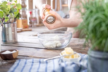In einer Küche mit Holzarbeitsplatte wird eine Glasschüssel mit Frischkäse mit einer Holz-Pfeffermühle gewürzt