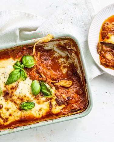 Ein Teller mit einer Portion Auberginen-Lasagne liegt neben der gefüllten Auflaufform mit knusprigem Käse und Tomatensauce.