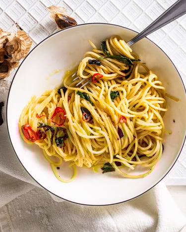 Auf einem weißen tiefen Teller sind Spaghetti in einer Knoblauchsoße angerichtet und von oben fotografiert. Um den Teller herum ist auf dem weißen Untergrund fermentierter Knoblauch verteilt