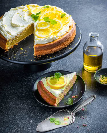 Ein angeschnittener Zitronenkuchen mit cremiger Sahne-Schmandhaube, die mit Zitronenscheiben und frischer Zitronenmelisse dekoriert ist, steht neben einer Flasche mit Olivenöl und einer Schale mit Zitronenabrieb auf einer Tortenplatte vor dunklem Hintergrund.