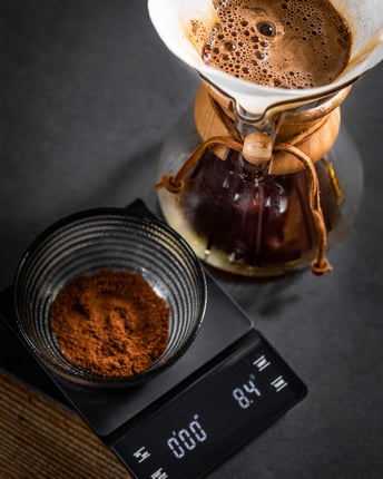 Feines Kaffeepulver, das in einer Glasschüssel auf einer Küchenwaage abgewogen wird, steht neben einer Chemex mit Handfliter, durch den frischer Kaffee läuft