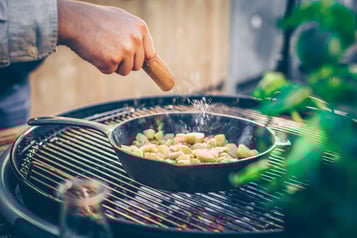 In einer gusseisernen Pfanne auf einem Grill sind gewürfelte Drillinge, die gerade mit Salz gewürzt werden. Im rechten Bildrand grüne Blätter außerhalb des Fokus.