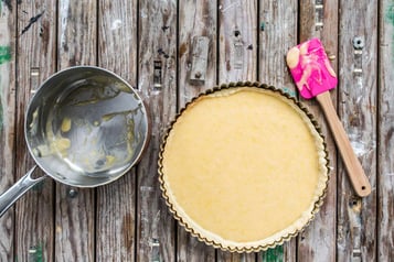 Tarteform mit Teig auf einem Holzuntergrund. Daneben ein Topf mit Stiel und ein pinker Teigschaber