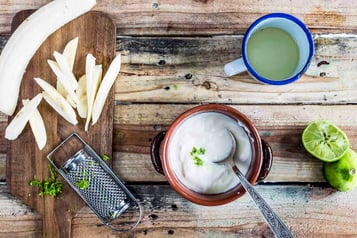 Joghurt mit Limettenabrieb in einer kleinen braunen Schüssel, links im Bild auf einem Holzbrett Banane in Stifte geschnitten