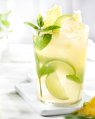 Ein großes Glas Limonade mit Limetten, Minze, Honigwaben und Eis gefüllt ist hinter einer Karaffe und vor einer Minz-Pflanze fokussiert.