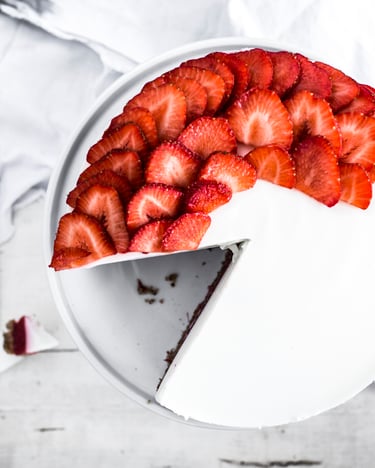 Aus einer weißen Torte, die zur Hälfte mit Erdbeerscheiben belegt ist, ist ein stück herausgeschnitten und liegt daneben auf einem weißen Brettchen,