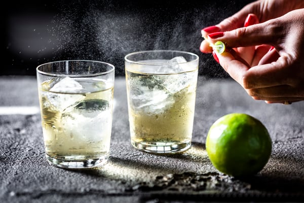 Zwei mit Eiswürfeln und einer hellgrünen Flüssigkeit gefüllte Gläser vor einem dunkelgrünen Untergrund neben einer Limette. Daneben sind weibliche Hände zu sehen, die eine Limettenzeste abreiben