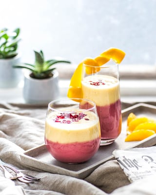 Zwei Gläser mit Two-Layer-Smoothie in rot und gelb auf Tablett auf beigem Leinentuch am Fenster