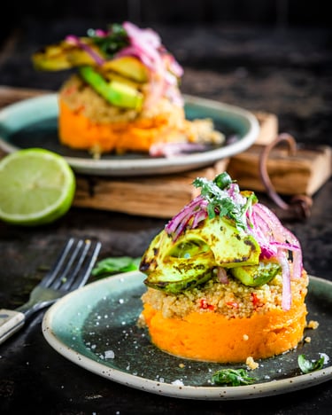 Süßkartoffel-Causa mit Quinoa und Avocado, das Schichtgericht ist angerichtet auf zwei farbigen Tellern und ein Teller ist auf einem Holzbrett drapiert, daneben eine halbe Limette, rote Zwiebeln und eine Gabel, fotografiert auf dunklem Untergrund.
