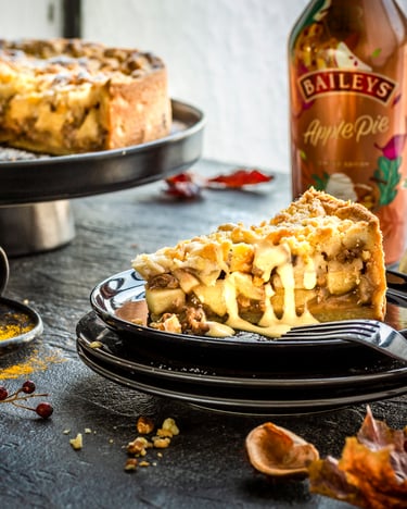 1 Stück Apple crumble Pie mit Baileys Vanillesauce  auf einem schwarzen Teller, dahinter auf einer Etagere der restliche Kuchen und eine Flasche Baileys Apple Pie.
