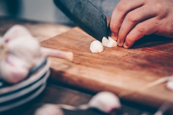 Knoblauch wird auf einem Holzbrett mit einem großen dunklen Messer geschnitten.