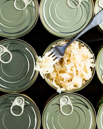 Eine offene Konservendose mit Sauerkraut steht inmitten geschlossener Konservendosen.