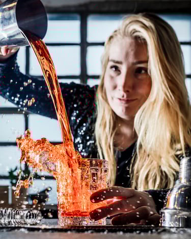 Eine junge Frau vor einer Barkulisse, die eine rote Flüssigkeit aus einem Cocktailshaker in ein Glas schüttet, sodass es spritzt
