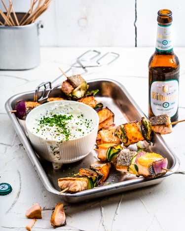 In einer Schale werden Grillspieße mit Lachs, Zucchini und roten Zwiebeln zu einem Sour-Cream-Dip und einer Flasche Bitburger Helles Lagerbier – glutenfrei  serviert.