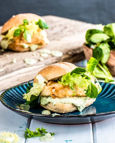 Belegte Fischbrötchen mit Salat und Sauce Tartare auf einem Teller und einem Holzbrett neben einer kleinen Schüssel Salat.