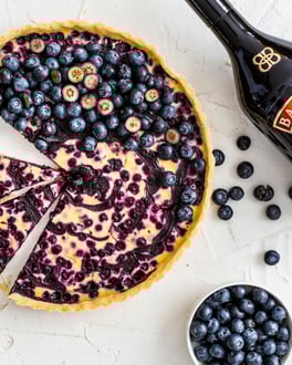 Tarte mit Blaubeeren dekoriert auf weißem Tisch mit Baileys The Original Irish Cream Flasche.
