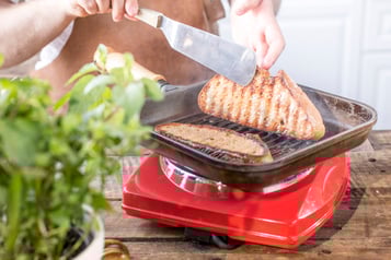 Zwei Graubrotscheiben wird in einer viereckigen Grillpfanne in Fett angeröstet. Eine Scheibe wird mit einer Hand und einer Palette gewendet. Die Pfanne steht auf einer roten Kochplatte, im linken Bildrand steht ein Topf mit Kräutern