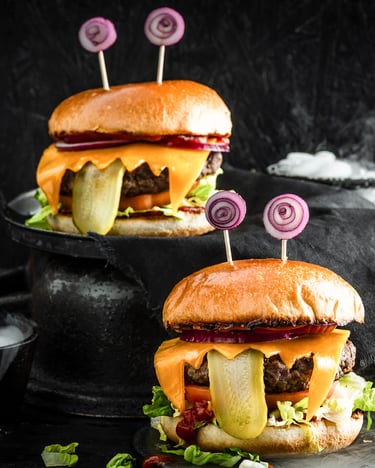 Zwei Burger, die durch hängende Gurkenzungen und aufgerollten Zwiebelaugen aussehen wie Monster. Links im Bild eine rauchende Schüssel. Der Bildhintergrund ist schwarz