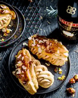 Eclairs gefüllt mit Espressocreme, glasiert mit Baileys und Haselnüssen garniert auf dunklen Desserttellern serviert neben einer Baileys Flasche.