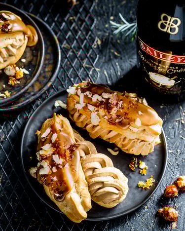 Eclairs gefüllt mit Espressocreme, glasiert mit Baileys und Haselnüssen garniert auf dunklen Desserttellern serviert neben einer Baileys Flasche.