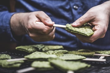 Ein grünes Waffelherz wird auf einen Holzstiel gesteckt. Darunter liegen weitere aufgespießte Waffeln