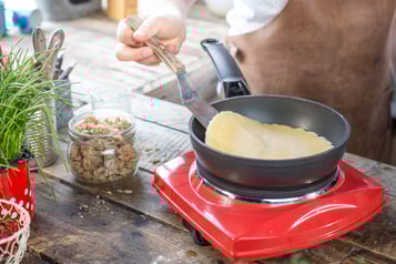 Auf einer roten Kochplatte in einer schwarzen Pfanne wird ein Pfannkuchen gebraten. Im Bildrand stehen Kräuter, Erdbeeren und ein Glas mit Müsli