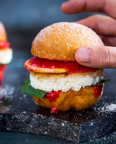 Vor schwarzem Hintergrund sind zwei süße Burger mit Brötchen aus Berlinern und einem Patty aus Milchreis. Eine Han greift von rechts nach dem Brötchendeckel des rechten Burgers
