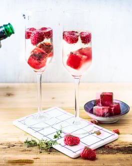 2 Gläser Sekt gefüllt mit Himbeer Eiswürfeln auf einem Holztisch