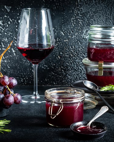 vor schwarzem Hintergrund stehen mehrere Gläser mit Trauben-Konfitüre und ein Glas Rotwein