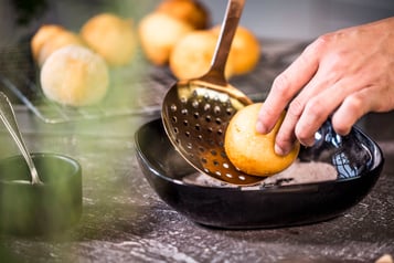 Ein Quarkbällchen wird mit Hilfe einer Schaumkelle und einer Hand in einer Zimt-Zucker-Mischung gewälzt. Im Hintergrund weitere Quarkbällchen auf einem Küchengitter