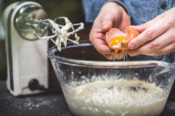 Ein Ei wird über einer großen Glasschüssel mit Waffelteig getrennt. Daneben steht ein Handrührgerät mit Teigresten an den Besen.