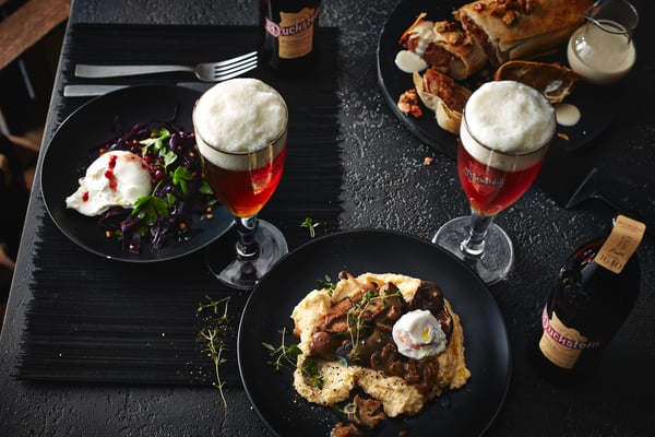 Ein 3-Gänge-Menü bestehend aus einer Burrata-Vorspeise, einer Parmesan-Pilz-Polenta als Hauptspeise und einem Dessert aus Apfelstrudel, steht neben rötlich schimmernden Biergläsern mit Duckstein Rotblond auf einem dunkel gedeckten Festtisch