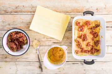 Lasagne mit gewürztem Hackfleisch in weiße Auflaufform geschichtet, daneben Lasagneplatten und zwei weiße Schüsseln mit Hackfleisch und Kichererbsensauce.