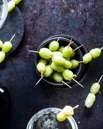 Gefrorene grüne Trauben an Zahnstochern stehen in Schälchen vor dunklem Hintergrund neben einer Flasche Prosecco und einem Glas Wodka.