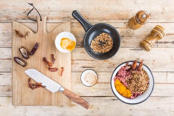 Datteln in Streifen geschnitten auf einem Holzbrett, daneben ein großes Küchenmesser. Rechts im Bild geröstete Mandeln in einer kleinen Pfanne und Hackfleisch mit Gewürzen in einer Schüssel.
