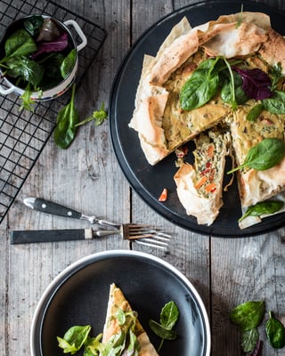 Auf einem Holzuntergrund steht ein schwarzer Teller mit einer Yufka-Quiche, daneben Messer und Gabel und ein weiteres Stück Quiche
