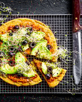 Socca Pizza mit Avocado und Oliven, mit Tomatengrund. Ein Achtel ist angeschnitten, Pizza wird von oben fotografiert und liegt auf einem Gitter.