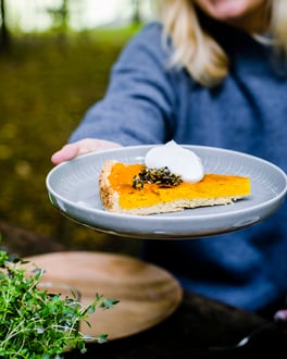Die Kürbis-Tarte ist knallorange und liegt mit einem dicken Klecks Sahne und Krokant auf einem hellblauen Teller, der von einer Frau in die Kamera gehalten wird.