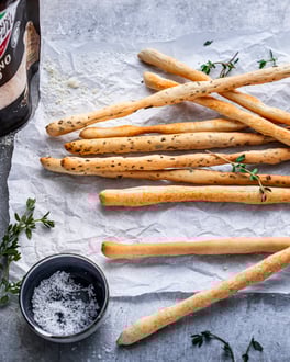 Grissini-Stangen auf weißem Backpapier mit Salz, Thymian, Olivenöl und Parmesan auf hellgrauem Betonuntergrund