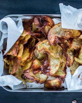 Knusprige, goldbraune, selbstgemachte Kartoffelchips auf Butterbrotpapier in glänzender Lunchbox, von oben fotografiert.