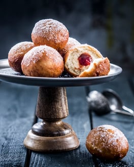 Mini-Berliner mit Erdbeerkonfitüre auf einem Serviertablett vor dunklem Hintergrund. Im Hintergrund liegt in der Unschärfe ein offenes Teesieb.