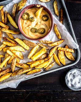 Pommes auf einem Backblech mit einer Schale Käse-Dip auf weißem Backpapier von oben