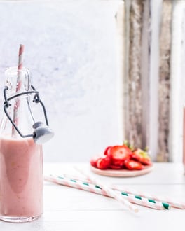 Drei kleine Glasflaschen mit rosa Erdbeer-Bananen-Smoothie und Papierstrohhalmen auf weißer Fensterbank