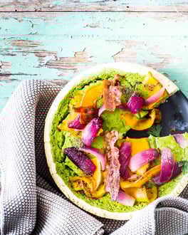 Grüne Quiche mit Kürbis und Speck, aus der ein Stück herausgeschnitten ist.