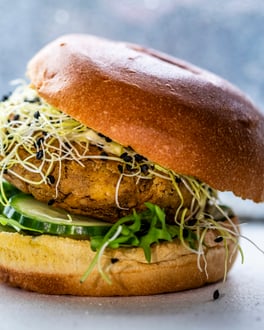 Burger Bun mit Gurken, Rucola und Knoblauchsprossen, außerdem belegt mit einem dicken Patty, der Deckel ist schräg aufgesetzt, fotografiert frontal vor einem hellen Fenster.