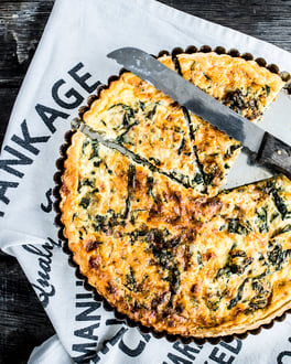 Mit einer goldenen Kruste zwischen der grüne Mangoldblätter durchscheinen steht die Mangold Quiche auf einem mit Text bedruckten Küchentuch. Ein Stück ist herausgeschnitten und darauf liegt ein großes Messer.